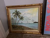 Photo of framed tropical beach painting showing palm trees, ocean, birds, and a golden bamboo-style frame.