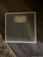 Square mechanical weight scale with analog dial showing weight measurement in kg.