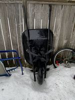 A large black metal wheelbarrow with single front tire leaning against a wooden fence on snow-covered ground, surrounded by other items such as a bicycle wheel and a small blue frame.