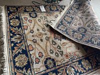 Full view of the rug partially rolled showing the navy blue floral border and beige background with floral motifs.