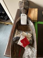 African carved wood spoon with elongated neck and woman's head, containing various items including pill box packaging and plastic utensils.