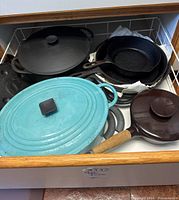 Drawer holding multiple cast iron items including turquoise Le Creuset Dutch oven with lid, three black cast iron skillets, and a brown pan with wooden handle.