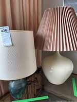 Two ceramic base table lamps side by side, one with green patterned base and white shade, the other with cream base and pleated beige shade.