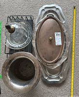 Top down view of all items showing the metal rack, glass butter dish with wooden knob lid, tarnished silver bowl, large oval silver-tone platter with decorative edges, smaller silver-tone platter with copper lid wrapped in plastic.