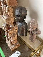 Overview of three sculptures on glass tabletop including tall multi-faced rider, dark wood bust, pink sandstone figure