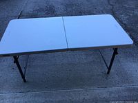 Full view of white rectangular folding table extended with black adjustable legs on concrete floor.