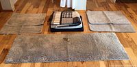 Top view of all items laid out on wooden floor: two folded blankets centrally stacked, flanked by four beige bath mats including one long medium bath mat in front
