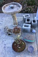 Full view of bird bath, frog planter with plants, cinder blocks, painted stone, rabbit ornament, sunflower dish, and a black round planter with dry plant