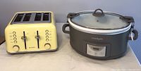 Light yellow 4-slice toaster with front panel controls and buttons, next to a large gray crockpot with glass lid and side handles, shown on a flat surface.