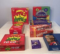 Overview shot showing six boxed games: Apples to Apples (regular and travel versions), two editions of Cranium, Yahtzee, Magnetic Sculpture kit, and a small wooden puzzle block set in a blue drawer-style box.