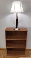 Small wooden bookcase with three shelves against a wall with a lamp placed on top.