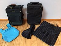 Front view of two rolling suitcases (green Ricardo Tiburon and black), turquoise backpack cooler, black crossbody bag and additional bags on wooden floor