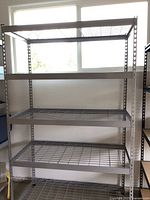 Front view of the metal shelving unit in front of a window showing four wire grid shelves and perforated uprights.