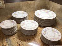 Multiple stacks of plates of various sizes and styles laid out on granite countertop, showing floral pattern and gold trim.