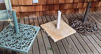 Three umbrella stands on wooden deck: one blue-green wrought iron with square ornate base, one brown wrought iron with octagonal ornate base, one polished marble with simple square base.
