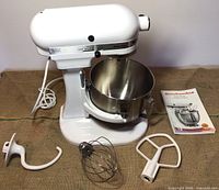 White tilt-head stand mixer with stainless steel bowl, three attachments, and manual arranged on burlap.