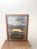 Framed print wall art photographed against a white wall showing the whole piece including the wooden frame and the printed inspirational quote over a sunset coastal image.