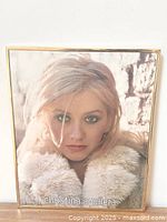 Full view of the framed Christina Aguilera poster mounted on a white wall, showing the face and fur collar with gold-toned frame and some frame wear.