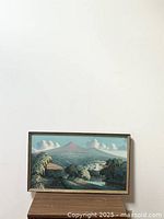 Frontal view of vintage oil painting in a thin frame showing a landscape with a mountain and greenery under cloudy sky.
