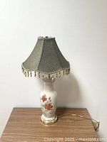 Full view of the vintage porcelain lamp on a wooden surface showing the floral painted base and gray striped fabric lampshade with beaded fringe.