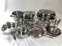 Overview of all stainless steel pots, pans, lids and roasting pans on white backdrop