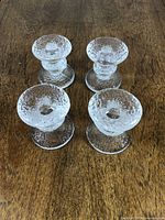 Four clear textured glass candlestick holders shown from an angle, all grouped together on a wooden surface.