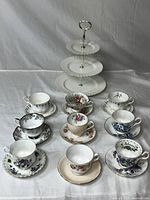 Top down view of all nine tea cups and saucers arranged in front of the three tier cake stand, showing assorted floral patterns and colors on the tea cups and saucers.