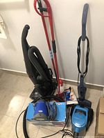 Multiple cleaning appliances including 3 upright vacuums, 2 red handle cleaning tools, and a blue Steam Zapper cleaner on its box, showing cords and hoses