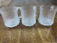 Three clear textured glass beer mugs arranged side by side on wooden table.