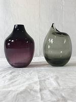 Two glass vases, a purple one and a grey asymmetrical one, displayed side by side on a white cloth.
