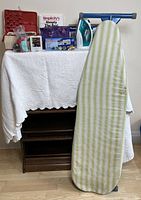 Wide shot showing the ironing board leaning against a fabric-covered table holding the sewing box, iron, box, and sewing accessories.