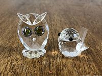 Front view of two Swarovski crystal figurines: an owl with green eyes and a bird with black eyes sitting on a wooden surface.