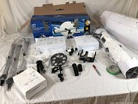 Disassembled telescope components laid out including tripod parts, eyepieces, mounting hardware, and instruction box in front of opened product box.