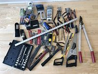 Top-down view of assorted hand tools spread out on light wooden floor, showing socket set, saws, hammers, pliers, cutters, measuring tape and smaller tools.