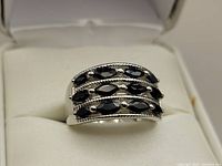 Front angle view of silver rhodium plated ring with three rows of dark blue marquise sapphires in white cushioned ring box.