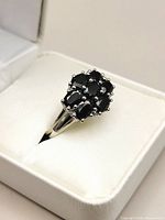 Front angled view of silver ring featuring dark blue sapphire stones cluster on top, ring placed on white cushioned ring holder inside white box.