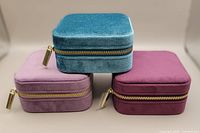 Three velvet square jewellery boxes stacked showing burgundy, teal, and light purple colors closed with gold zipper pulls