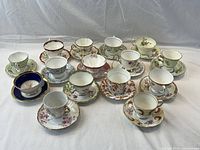 Photo showing 15 tea cups and matching saucers arranged in rows, displaying variety of floral and decorative patterns with gold trim