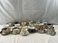 Full view of all 18 teacups with matching saucers arranged in two rows on white cloth, showing variety in patterns and colors.