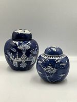 Two blue and white porcelain ginger jars with lids placed on a flat surface with neutral background. Larger jar decorated with intricate white floral and ornamental designs on a blue background, smaller jar also blue with a white floral pattern.