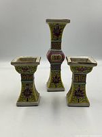Three Chinese Famille Rose porcelain candle holders with square tops and bases, decorated in yellow, pink, green, and blue. Two shorter identical holders flank a taller one with a bulbous midsection.
