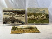 Three oil paintings on board displayed on white cloth: snowy landscape, blossoming orchard, haystacks field
