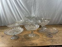 9 assorted vintage pressed glass compotes arranged on wooden surface, displaying various patterns and sizes
