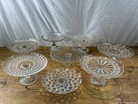 Eight pressed glass cake stands arranged on wooden surface showing variety of patterns and sizes