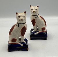 Front view of the pair of white porcelain cats with brown patches, sitting on blue pillows, showing both cats and their painted facial details and collars.