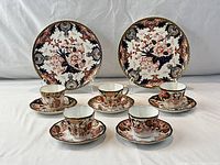 Set showing 2 plates standing in back, 4 cups on 4 saucers arranged in front with intricate floral and vine pattern in dark blue, burnt orange, black, and gold trim.