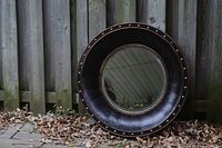 Round mirror with wide dark frame and metal inlays, leaning against wooden fence on ground with leaves around.