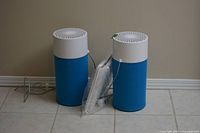 Two Blueair compact air purifiers alongside one extra sealed replacement filter, positioned on tiled floor near wall outlet.