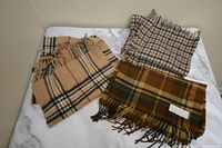 Three scarves laid on a white and gray marble surface, showing varying plaid and check patterns with fringed edges.