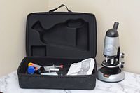 Silver and black children's microscope beside open black carrying case on white marble surface